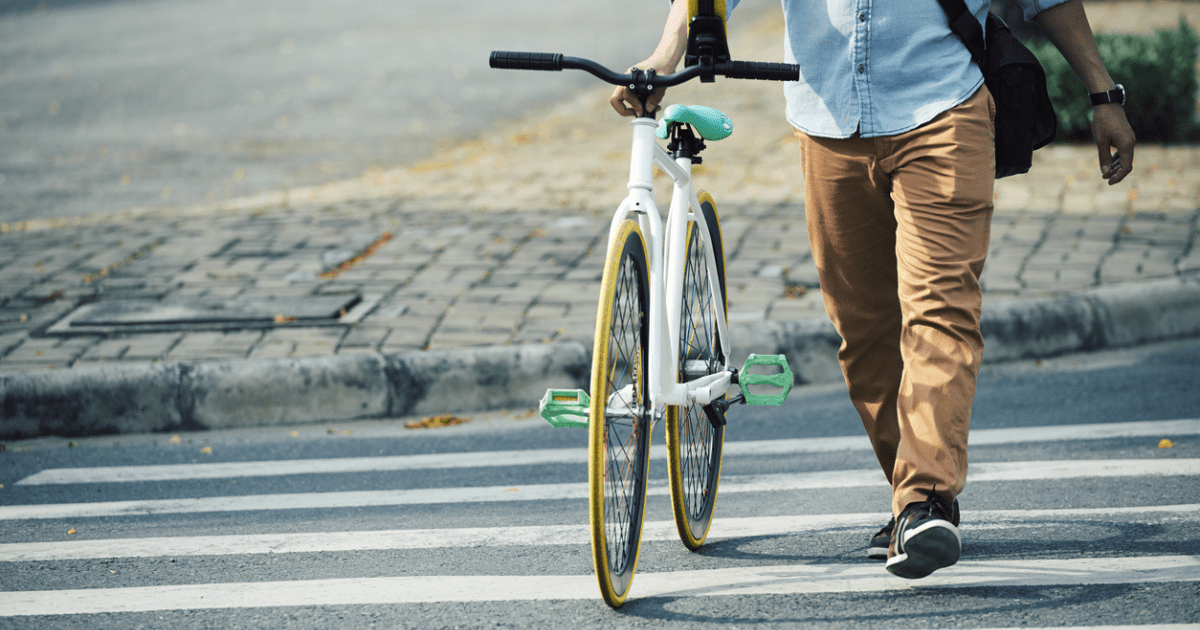 A man walking his bike across a crosswalk in the city carrying his urban edc backpack.